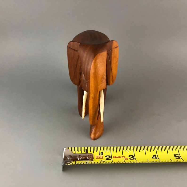 May include: A wooden elephant figurine with carved details and ivory-colored tusks. The elephant is standing on a gray surface with a yellow measuring tape in the foreground.