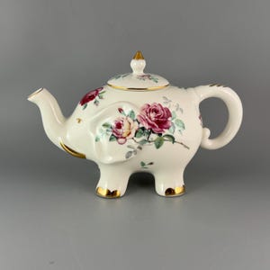 May include: A cream-colored ceramic teapot shaped like an elephant, featuring a floral pattern of pink and red roses. The teapot includes a matching lid with a gold-painted finial, gold trim on the spout, handle, and feet, and a curved trunk.