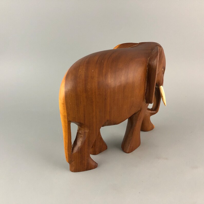 May include: A wooden elephant figurine with a light brown color and a carved ivory tusk. The elephant is standing with its legs slightly bent and its trunk raised.