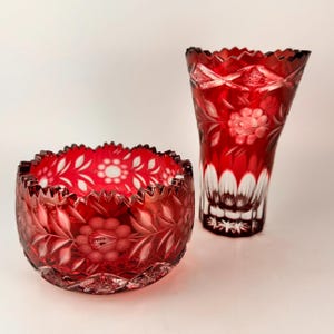 May include: A set of two red crystal bowls with intricate white floral designs. The bowl on the left is round and the bowl on the right is a vase.
