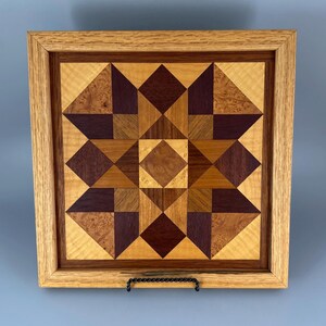 May include: A square wooden wall art piece featuring a geometric star pattern made from inlaid wood segments in shades of light tan, medium brown, and deep purple. The artwork is enclosed in a light-coloured wooden frame with a visible grain.