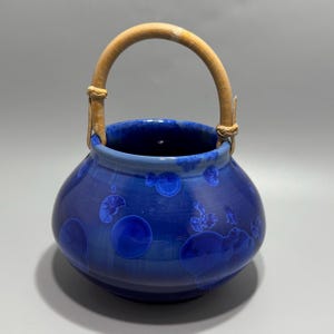 May include: A round ceramic vessel featuring a deep blue crystalline glaze with lighter blue circular patterns. The pot has a wide opening and a curved, light-colored bamboo handle attached to the sides with woven fiber bindings.