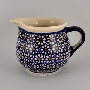 May include: A ceramic creamer with a dark blue floral pattern. The creamer has a white interior and a dark blue handle. The floral pattern consists of white and orange-brown flower-like shapes on a dark blue background.