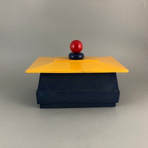 May include: A wooden box with a yellow lid and a blue base. The lid has a red ball on top of a blue ring.