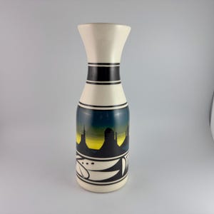 May include: A white ceramic vase with a black and yellow design depicting a desert landscape. The vase has a narrow neck and a wide base.