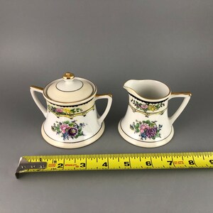 May include: A vintage porcelain sugar bowl and creamer set with a floral design. The set is white with gold accents and features a yellow, pink, and purple floral pattern.