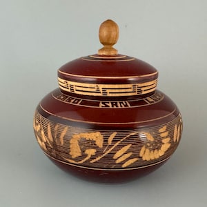 Peut inclure: Un pot rond en bois fait à la main avec un couvercle. Le pot est de couleur brun rougeâtre foncé avec des motifs gravés clairs et les mots "SAN LUIS" visibles. Le couvercle a un petit bouton en bois.