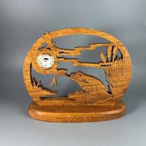 May include: A wooden clock with a frog design. The clock is shaped like an oval and has a carved frog on the bottom. The clock face is round and has a black background with white numbers. The clock is on a wooden base.