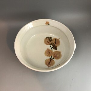 Könnte beinhalten: Eine weiße Keramikschüssel mit braunem Blattdesign. Die Schüssel hat zwei Griffe an den Seiten.
