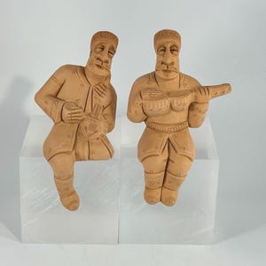 Puede incluir: Dos figuritas de terracota de músicos, uno tocando un instrumento de cuerda y el otro un instrumento de viento. Las figuritas están sentadas sobre pequeños pedestales blancos.