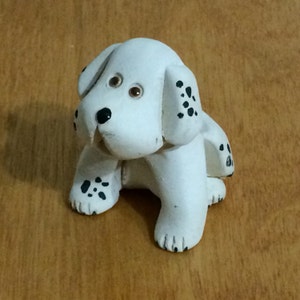 May include: A small white ceramic figurine of a Dalmatian puppy with black spots. The puppy is sitting with its head tilted to the side and its eyes closed.