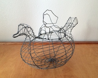 May include: A wire teapot shaped like a bird, with a rounded body and a spout that curves up like a neck. The teapot is made of thin wire and has a delicate, airy appearance.