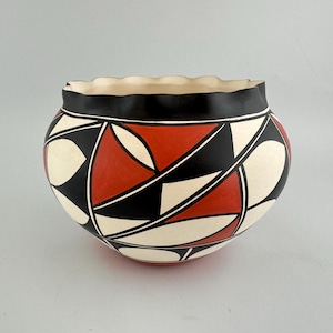 May include: A round ceramic bowl with a scalloped rim. The bowl features a geometric design in red, black, and cream colors. The rim is black, and the interior is cream-colored. The bowl is likely handmade and has a traditional aesthetic.