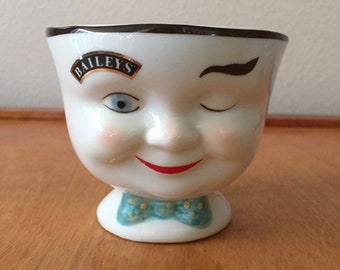 May include: A white ceramic egg cup with a smiling face design. The cup features a winking eye, rosy cheeks, and a red smiling mouth. It has a green bow tie with yellow dots and the "Baileys" logo.