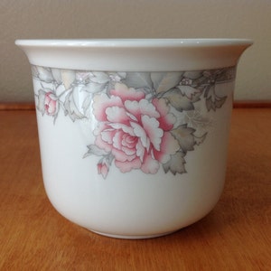May include: A white ceramic planter with a floral design. The planter features a large pink peony flower with gray and green leaves. The rim of the planter has a decorative border. The planter is cylindrical in shape.