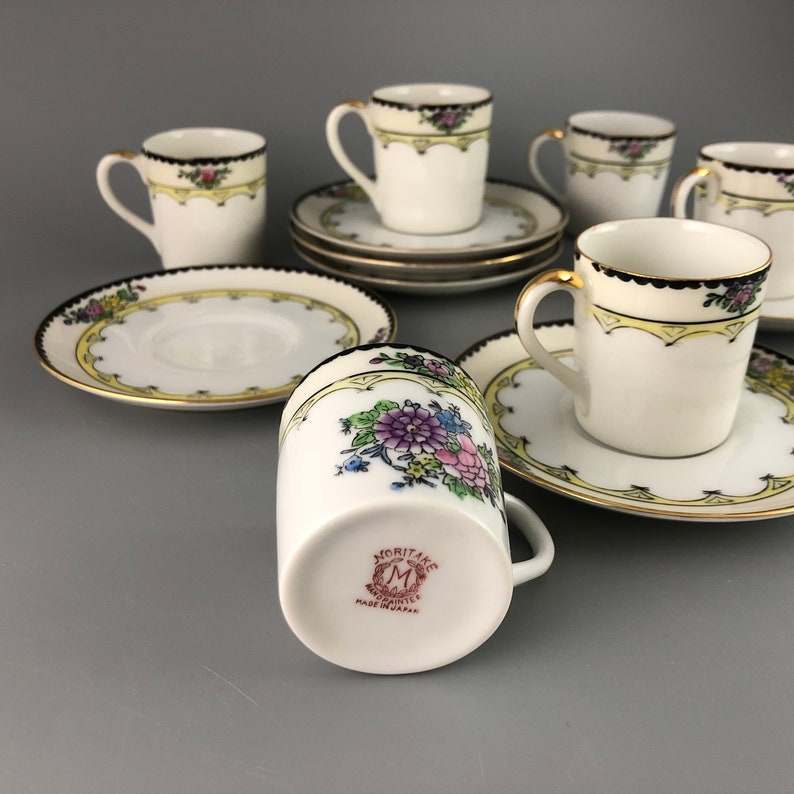 May include: Set of six vintage Japanese teacups and saucers with floral designs. The cups and saucers are white with gold trim and feature a black band around the rim. The cups have a floral design with pink, purple, and blue flowers. The saucers have a similar floral design with a yellow border. The cups and saucers are marked "Moritane" and "Made in Japan".
