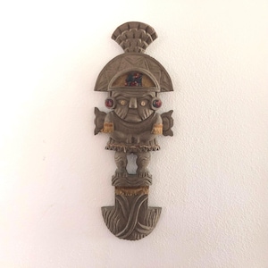 May include: A carved wooden wall hanging depicting a stylized figure with a large headdress and ornate details. The figure has red accents and a detailed face. The piece is a light brown color and appears to be a decorative item.