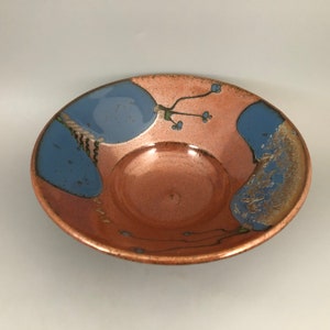 May include: A ceramic bowl with a copper glaze and blue accents. The bowl has a unique design with a raised center and a rim that curves inward.