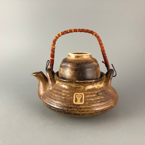 May include: A brown ceramic teapot with a lid and a woven handle. The teapot has a decorative design on the side with a stylized flower.