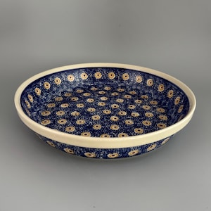May include: A round, ceramic serving dish with a cream-colored rim and a dark blue interior. The interior is decorated with a repeating pattern of small, yellow and brown floral designs. The dish is ideal for serving food.