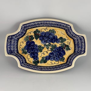 May include: An oval ceramic serving platter with handles, featuring a central yellow background decorated with three clusters of blue grapes and green leaves, surrounded by a wide border with a repeating dark blue geometric pattern.