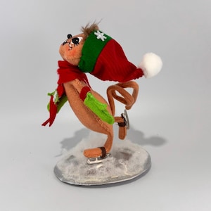 May include: A brown and tan felt squirrel wearing a red and green hat with a white snowflake, green mittens, and a red scarf, ice skating on a silver base.