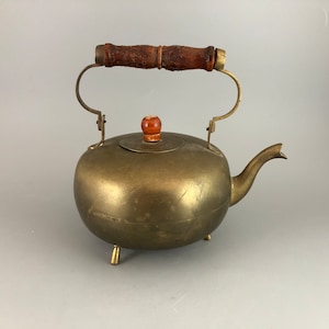 May include: A brass teapot with a wooden handle and lid knob. The teapot has three legs and a rounded body.