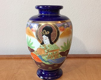 May include: A cobalt blue ceramic vase with a hand-painted design of a woman in a colorful robe holding a mirror. The vase has gold accents and a white background.