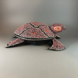 Pode incluir: Uma estatueta de tartaruga preta com uma carapaça coberta de pontos rosa e brancos, e alguns pontos laranja. A cabeça e as barbatanas da tartaruga também são pretas com detalhes brancos e laranja. Os olhos são delineados em laranja.