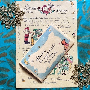 Puede incluir: Una carta navideña festiva dirigida a Daniel, con ilustraciones de Papá Noel y elfos. La carta va acompañada de una pequeña tarjeta con el texto "Daniel, la estrella más brillante en una habitación llena de estrellas". Adornos de copos de nieve dorados adornan la escena.