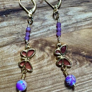 May include: A pair of gold-tone leverback earrings featuring a vertical arrangement of three small purple faceted beads, a gold-tone butterfly charm with translucent reddish-brown wings, and a single round purple and white marbled bead at the base.