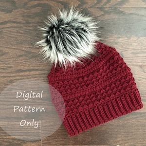 May include: A burgundy crochet beanie with a black and white faux fur pom pom. The beanie has a textured stitch pattern. Digital Pattern Only.