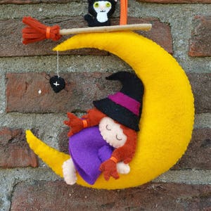 Op de afbeelding: Een vilt Halloween-decoratie met een slapende heks op een gele halve maan. Een zwarte kat met witte markeringen zit op een bezem boven de maan. Een spin hangt aan een touwtje boven de heks.