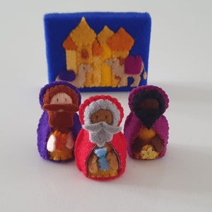 Op de afbeelding: Drie vingerpoppen van vilt die de drie wijzen uit het oosten voorstellen, elk met een gewaad in een andere kleur: paars, rood en bordeauxrood. De poppen staan voor een vilt achtergrond die een kasteel met een kameel voorstelt.
