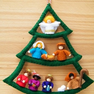Puede incluir: Un adorno de árbol de Navidad de fieltro con un árbol de fieltro verde y figuras de fieltro que representan la escena de la Natividad. Las figuras incluyen a María, José, el niño Jesús, un ángel, un pastor y una oveja.
