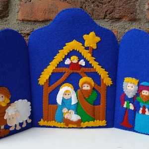 May include: A felt Christmas nativity scene with a blue background. The scene includes Mary, Joseph, baby Jesus, a shepherd, a sheep, and the three wise men. The scene is decorated with a yellow star and a yellow straw roof.