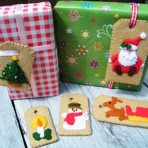Op de afbeelding: Vijf vilt kerstcadeau-etiketten met verschillende feestthema's. De etiketten zijn beige met rode, groene, witte en gele accenten. De thema's omvatten een kerstboom, een sneeuwpop, een rendier dat een slee trekt, een kaars en de kerstman.
