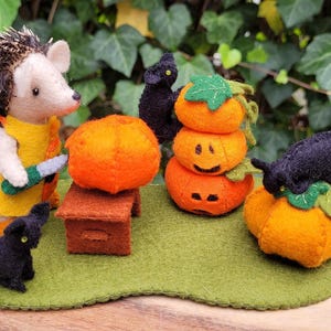 Può includere: Un riccio di feltro con un vestito giallo e un coltello taglia una zucca arancione di feltro. Due gatti neri di feltro sono seduti vicino a una pila di tre zucche arancioni di feltro con facce di zucca.