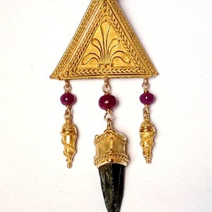 May include: A gold pendant necklace with a triangular pendant featuring a stylized floral design. The pendant is attached to three chains with red beads and gold accents. The pendant is also attached to a dark brown cord.