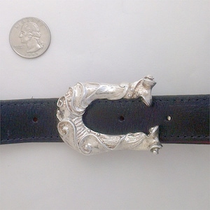May include: A silver belt buckle with an ornate, abstract design. The buckle is attached to a black leather belt.