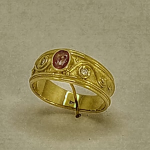 Puede incluir: Un anillo de banda de oro con una piedra preciosa rosa y dos piedras blancas más pequeñas. La banda está decorada con un diseño de espiral.