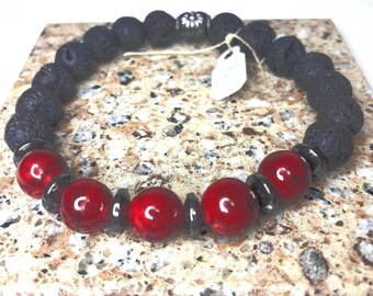 May include: A black lava rock bracelet with red beads and a silver charm. The bracelet is on a light brown surface.