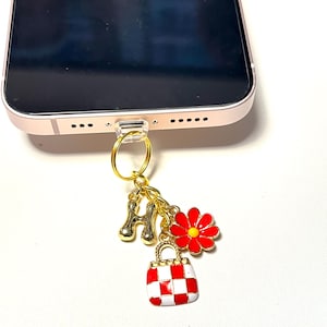 May include: A gold-colored phone charm with a clear plastic loop attached to a phone. The charm features a gold letter "H", a red flower with a yellow center, and a red and white checkered handbag.