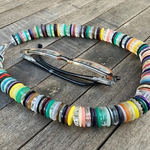 Puede incluir: Un collar colorido hecho de cuentas redondas translúcidas de varios tamaños en tonos rosa, verde, amarillo y morado. El collar descansa sobre una superficie de madera, con un par de gafas dentro.