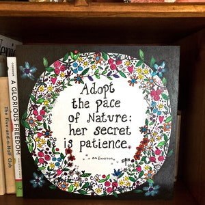 May include: A black canvas print featuring a colourful floral wreath surrounding the text "Adopt the pace of Nature; her secret is patience." The quote is attributed to Emerson.