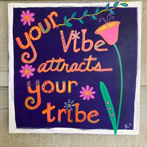 May include: A purple background with white trim features a hand-painted sign with the text "Your vibe attracts your tribe" in orange and pink lettering. The sign is decorated with flowers and vines in various colors.