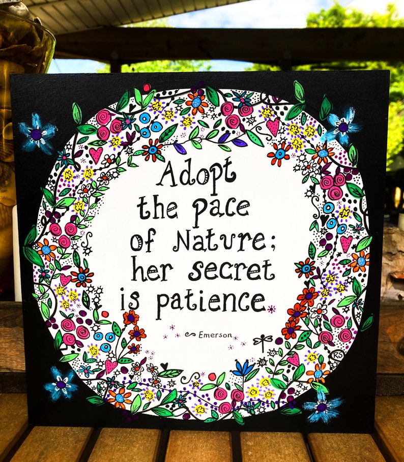 May include: A black canvas with a white circle surrounded by a colourful floral wreath. The quote "Adopt the pace of nature; her secret is patience." is written in black script inside the circle. The quote is attributed to Emerson.