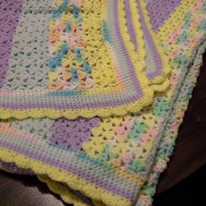 May include: A crocheted baby blanket in shades of purple, blue, yellow, and pink. The blanket has a scalloped edge and is made with a variety of stitches.