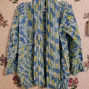 May include: A handmade, open-front crochet cardigan in shades of blue, green, and yellow. The long-sleeved sweater features a textured stitch pattern and is displayed on a hanger against a floral wallpaper background. The yarn appears to be a soft, bulky weight.