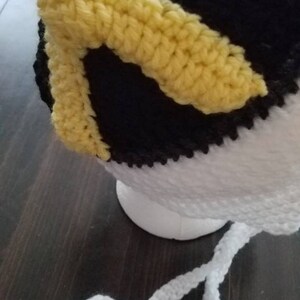 May include: A crocheted hat with black, white, and yellow stripes. The hat has a white crocheted tie.